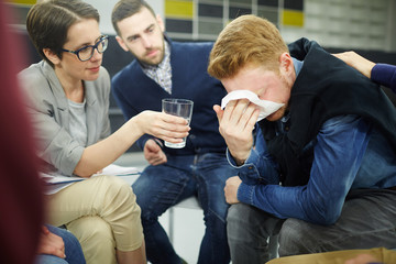 Suffering man covering his face by handkerchief while his counselor giving him glass of water