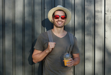 Outdoor picture of young European male standing in sun hat and sunglasses with gray wooden fence...