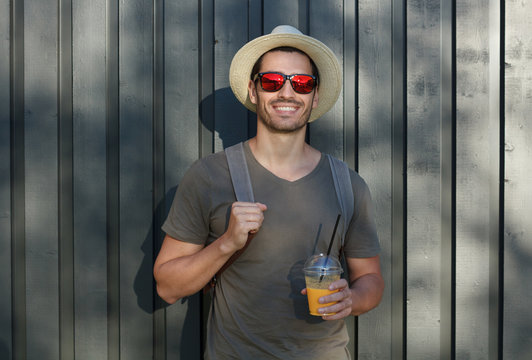 Outdoor picture of young European male standing in sun hat and sunglasses with gray wooden fence behind his back, holding strap of backpack and cup of drink, willing to continue walking around