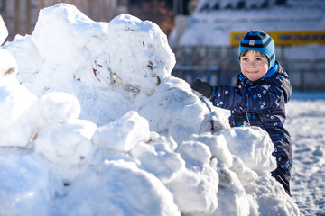 Funny little kid boy in colorful clothes playing outdoors during snowfall. Active leisure with children in winter on cold snowy days. Happy child having fun and snow