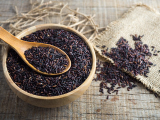 Close up rice berry(brown rice, Thai black jasmine rice or raw purple/red rice) in a wooden bowl, spoon and sack on wooden background.