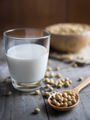 A glass of soy milk, soy beans in a wooden spoon and wooden bowl on wooden board.