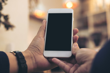 Mockup image of woman's hands holding and using white mobile phone with blank black screen in vintage cafe