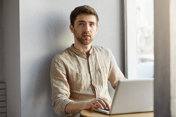 Handsome unshaven guy with dark hair working in coworking office near window, looking aside with thoughtful expression, trying to remember things he need to do.