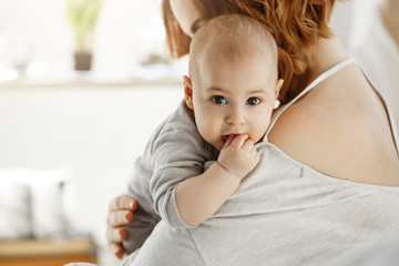 Fototapeta premium Portrait of sweet little child looking at camera with big grey eyes and putting hand in mouth on mother shoulder. Mom hug her child with love. Family concept.