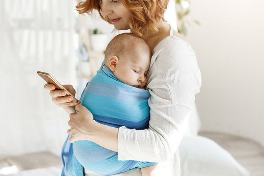 Tiny baby boy having pleasant dreams in baby sling while mother rests and looking through social networks on smartphone. Family, lifestyle concept.