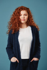 Portrait of beautiful ginger student girl with wavy hair and freckles holding hands in pockets, looking in camera with satisfied look. Vertical shot.