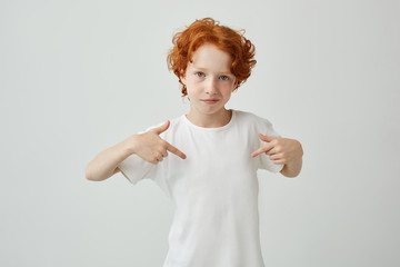 Close up of red haired cute boy with freckles pointing with fingers on white t shirt with serious...