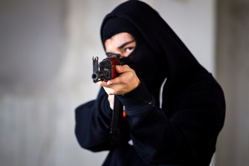 Unidentified man in black suit with face masked is aiming assault rifle to the target close up.