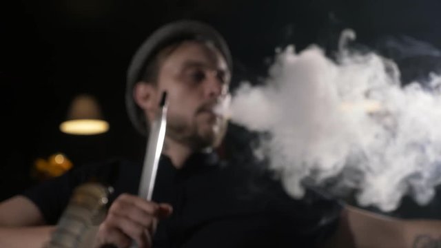 Handsome young stylish men in hat smoking shisha at dark cafe
