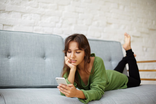Candid Shot Of Serious Young Housewife Of Hispanic Appearance Texting Husband On Smart Phone, Lying On Grey Sofa. Attractive Woman Having Rest At Home On Couch, Browsing Newsfeed Via Social Networks