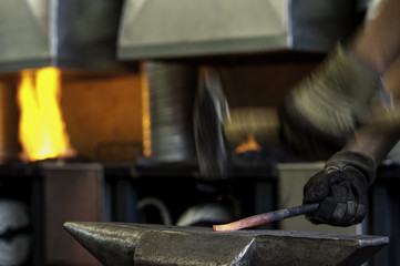 Hammer schlägt mit viel Schwung auf glühendes Eisen am Amboss, Feuer in der Esse unscharf im...