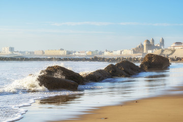 Cadiz city with the Ancient Cadiz Cathedral on the background. Big stones on the shore. The waves on the winter Atlantic Ocean. Spain