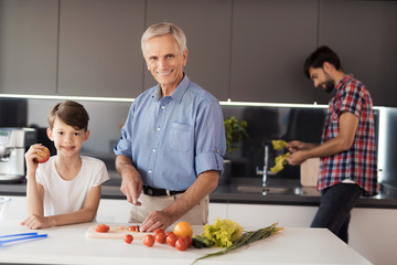 The old man and the boy are posing in the kitchen for making salad. The man behind them washes the lettuce leaves