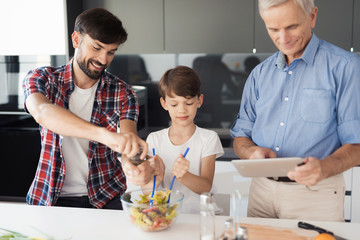 The boy and his father finish the salad for Thanksgiving. The old man stands next to them and looks at his gray tablet