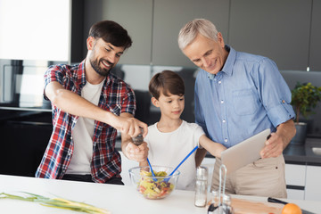 A man is preparing a salad. The boy and the old man stand side by side and look at something on the old man's tablet
