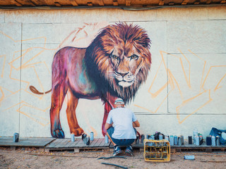 Graffiti Artist Painting On The Street Wall. Man with aerosol spray bottle near the wall. Young talented guy in protective mask drawing colorful lion.