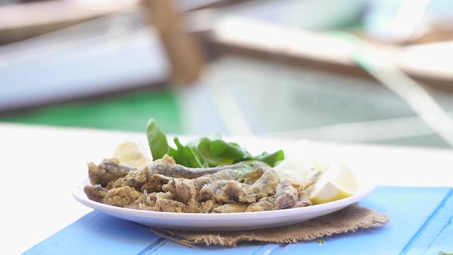 Turkish Hamsi Tava with lemon and arugula on blue table. Fried Anchovies by the sea at Foca Izmir. Copy space with sea and small boats background.