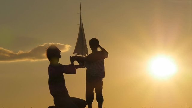 A father and a child hold a sailboat against the sky. Backlight. Silhouettes of people against the sky and sun. Model of a sailboat.