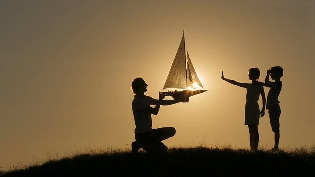 A model of a sailboat. A man is holding a ship model against the sky. The father is giving the ship to the child. Backlight. Silhouettes of people against the sky and the sun.