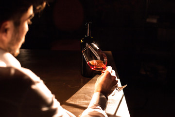 The handsome man is holsing a glass of red wine in his hand while sitting in the dark room.