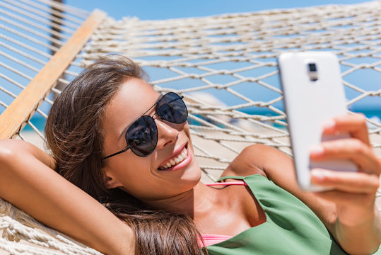 Vacation woman using smart phone taking smartphone selfie. Girl relaxing on beach hammock sun tanning in sunglasses smiling using cellphone to take pictures and text sms messages.