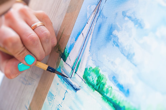 Close-up Young woman painter drawing watercolor and wooden brush on white paper seascape with a single-deck yacht