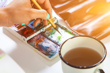 Close-up artist with bright nails dabs a brush in a pallet with a watercolor a thin wooden brush, on the table there is a leather cover with brushes and a mug of water