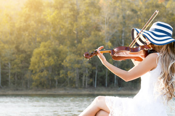 A woman / violinist wearing white dress and hat playing violin on riverside among sunlight feeling freedom © Nusara