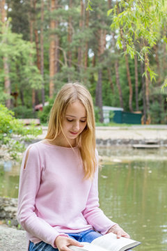 Cute romantic blond teenage-girl is reading a book sitting under the tree in the city park in the sunny weather. A pond surrounded by rocks with trees reflexion in the water is at the background.