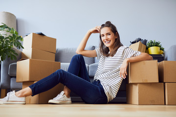 Cheerful young woman sitting in new apartment leaning on boxes after moving in