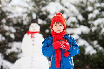 Happy beautiful child building snowman in garden, winter time