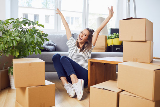 Ecstatic young woman sitting on floor in new apartment with boxes and raising arms in joy