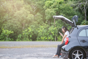 Naklejka premium Happy asian woman sitting on trunk of hatchback car in the park and using smartphone with forrest background