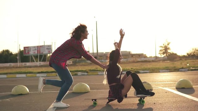 Side view of a woman with her hands raised up sitting on a longboard while her friend is pushing her behind and running during sunset. Enjoying life. Lens flare. Slowmotion shot