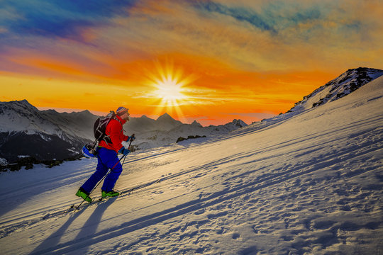 Mountaineer backcountry ski sunrise walking up along a snowy ridge with skis. In background cloudy sky and shiny sun and Swiss Alps.
