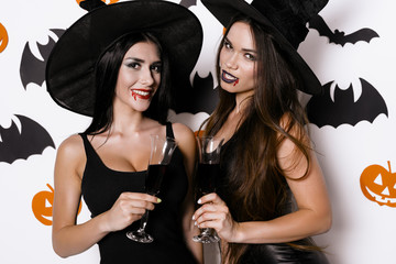 Two vampire witches are posing against a background of bats with glasses of blood in their hands