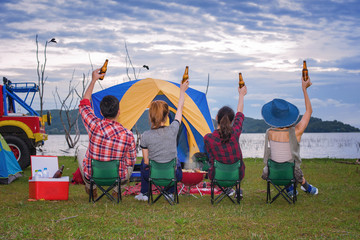 group of adventure tourist camping cheerful and enjoy together by take up bottle beer in the air, Barbecue camping at the lake in background