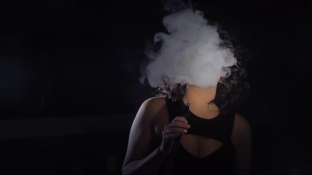 Horizontal glamour portrait. The woman with black hair is chilling by smoking the hookah and producing the thick smoke in the black studio.