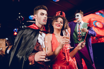A guy dressed as a vampire and a girl dressed as a demon posing with champagne glasses in their...