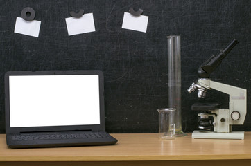 Teacher or student desk table. Education background. Education concept. Laptop with blank screen, microscope, flasks and note paper on blackboard (chalkboard) background. Chemistry or biology lesson.