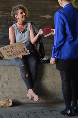 a punk woman sitting under a bridge with bottle of alcohol next by holding a sign and getting pleased a bible from a businesswoman