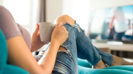 Beautiful young woman relaxing on the sofa at home, watching tv and enjoying coffee
