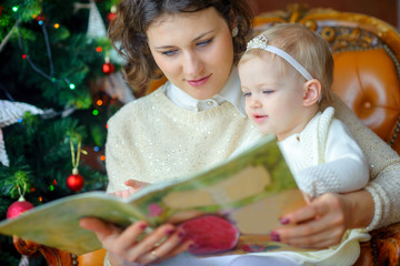 Mum reads fairy tales to a small daughter sitting near a festive Christmas tree.