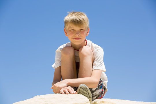 Young blonde boy sits on rock, look at camera and smiles