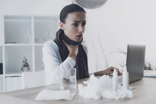 Sick woman sitting at her workplace in the office. She has a sore throat and she wore a scarf