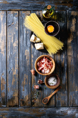Ingredients for traditional italian pasta alla carbonara. Uncooked spaghetti, pancetta bacon, parmesan cheese, egg yolk, salt, pepper in olive wood bowls over old plank background. Top view with space
