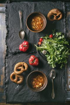 Clay bowls with vegetarian hot carrot tomato pea potato soup, served with fresh coriander, pretzels bread and tomatoes on textile napkin over old wooden plank table. Flat lay