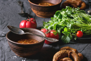 Clay bowls with vegetarian hot carrot tomato pea potato soup, served with fresh coriander, pretzels bread and tomatoes on textile napkin over old wooden plank table. Rustic style dinner