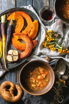 Fall holiday table decoration setting with bowls of hot carrot potato soup, baking pumpkin, carrot, garlic, pretzels bread, red wine, orange berries. Flat lay over wooden table